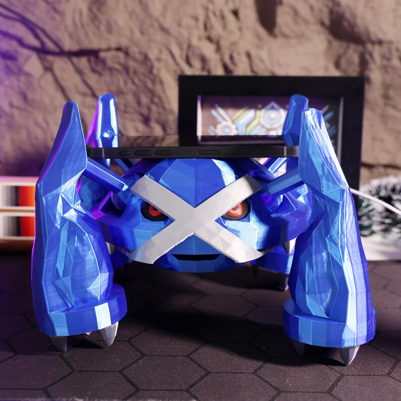 May include: A blue and white 3D printed phone stand shaped like a Pokemon character. The stand has a large, flat surface for holding a phone.