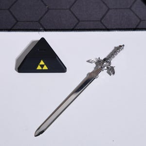 May include: A silver-colored Master Sword replica with a black triangular stand featuring the Triforce symbol. The sword has a detailed hilt and a serrated blade.