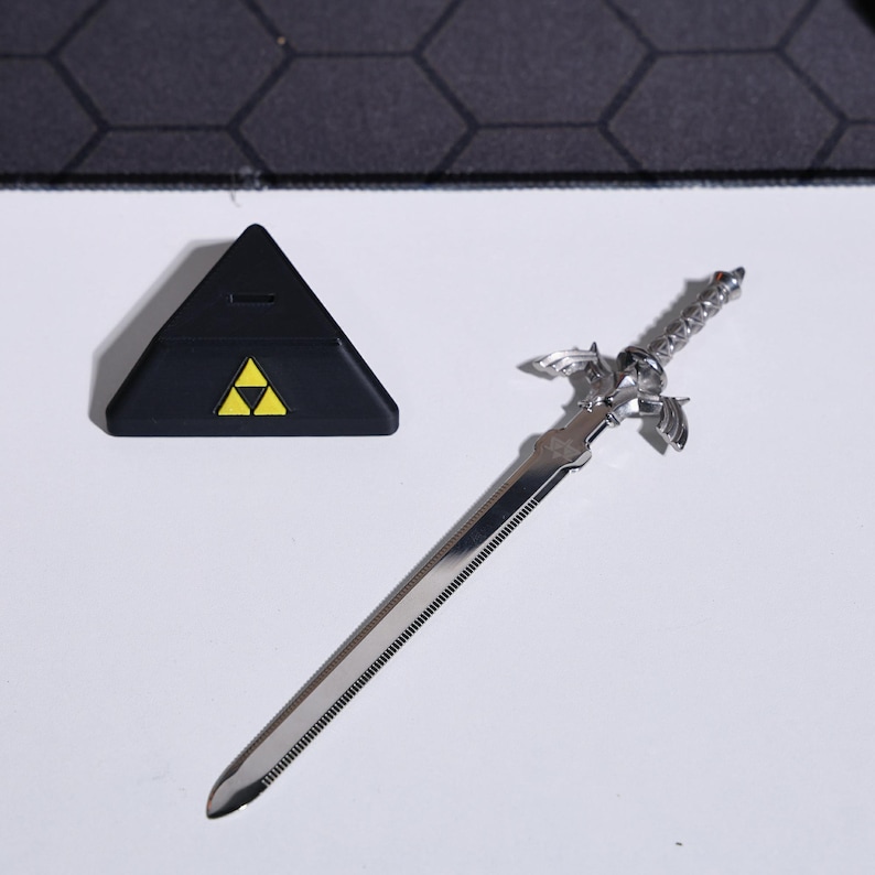 May include: A silver-colored Master Sword replica with a black triangular stand featuring the Triforce symbol. The sword has a detailed hilt and a serrated blade.