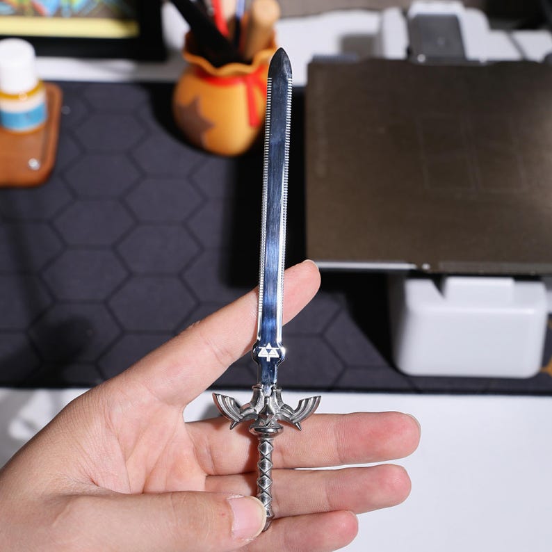 May include: A silver-colored Master Sword replica with a detailed hilt and a serrated blade. The sword is held in a hand.