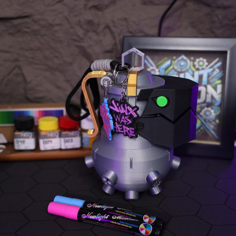 May include: A 3D printed gray and black Jinx grenade keychain with a purple and pink charm that says "Jinx Was Here".