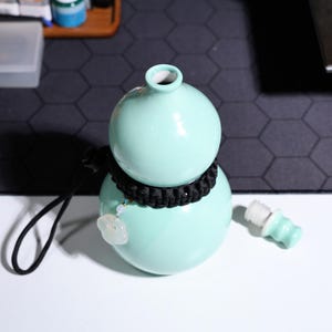 May include: A light blue ceramic gourd-shaped bottle with a black braided cord around the neck. The bottle has a small jade-colored charm attached to the cord. A separate light blue bottle stopper is lying on a white surface.