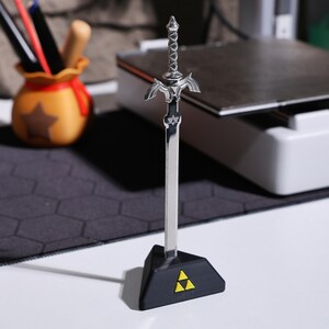 May include: A silver Master Sword replica with a black plastic stand featuring the Triforce symbol. The sword is on a black and gray textured surface.