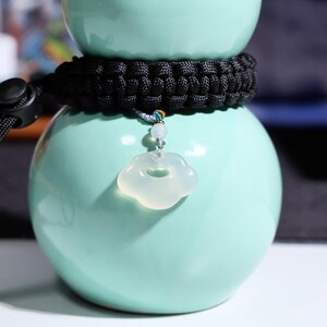 May include: A white jade pendant shaped like a cloud with a small hole in the center. The pendant is hanging from a black braided cord.