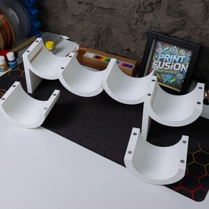 May include: White wall-mounted shelf unit with five curved compartments for displaying items. The shelf is made of white plastic and has a modern design. The shelf is assembled and ready to be mounted on a wall.
