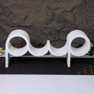 May include: White shelf unit shaped like the word "pup" with rounded edges and circular openings. The shelf unit is made of a smooth, white material and is designed to hold small items.