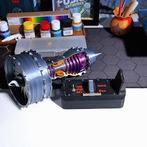 Op de afbeelding: Een gedetailleerd model van een straalmotor met een doorsnede van de interne componenten. Het model is gemaakt van grijs en zilver plastic met paarse en oranje accenten. De motor is gemonteerd op een zwarte basis met een bedieningspaneel met knoppen met de labels "FWD", "STOP", "REV", "Quick" en "Slow".