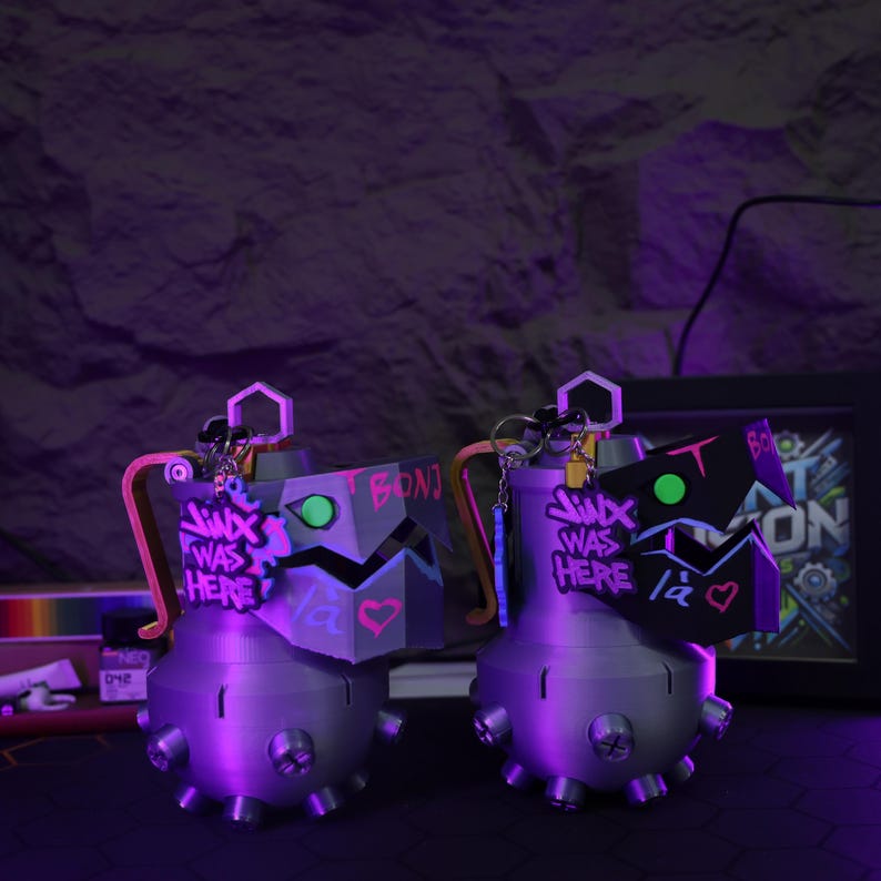 May include: Two gray, round, grenade-shaped keychains with black and green details. The keychains have a pink and purple graffiti design that says "Jinx was here" and "I BONJ".