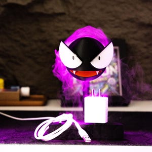 May include: A black and white cartoon character with a red mouth and white eyes, emitting purple vapor. The character is mounted on a white charger with a USB cable. The overall theme is a pop culture item.