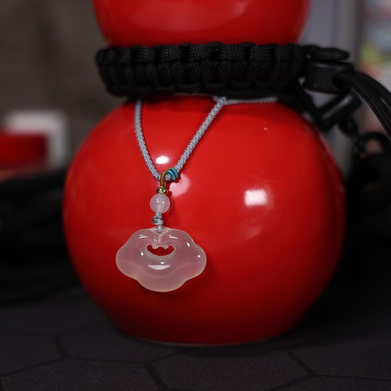 May include: A pink jade pendant shaped like a cloud hangs from a gray cord on a red ceramic vase. The pendant has a gold accent and a small blue bead.