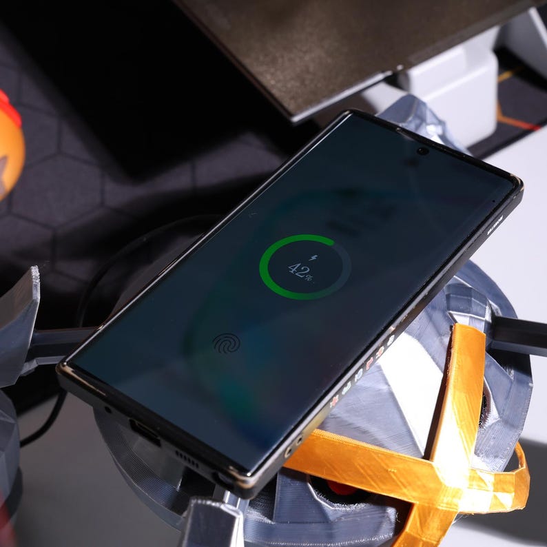 May include: A black smartphone with a fingerprint sensor is charging on a wireless charging pad. The phone's screen shows a green charging circle with the text "42%".