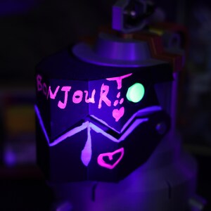 May include: A black, geometrically shaped object with a pink inscription that reads "Bonjour!" and a green glow. The object has two pink hearts drawn on it.