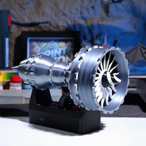 Op de afbeelding: Een 3D-geprint model van een straalmotor in zilver en wit op een zwarte basis. De motor heeft een ventilator met witte bladen en een zilveren behuizing.