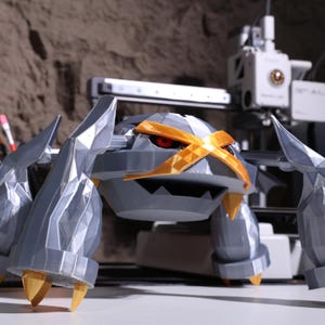 May include: A 3D printed model of a gray and gold Pokemon character. The character has a large, round head with a cross-shaped design on its forehead. The character has four legs and two arms, and its eyes are red.