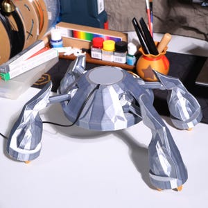 May include: A 3D printed, silver-colored, multi-legged robot with a round, flat top. The robot is sitting on a white surface.