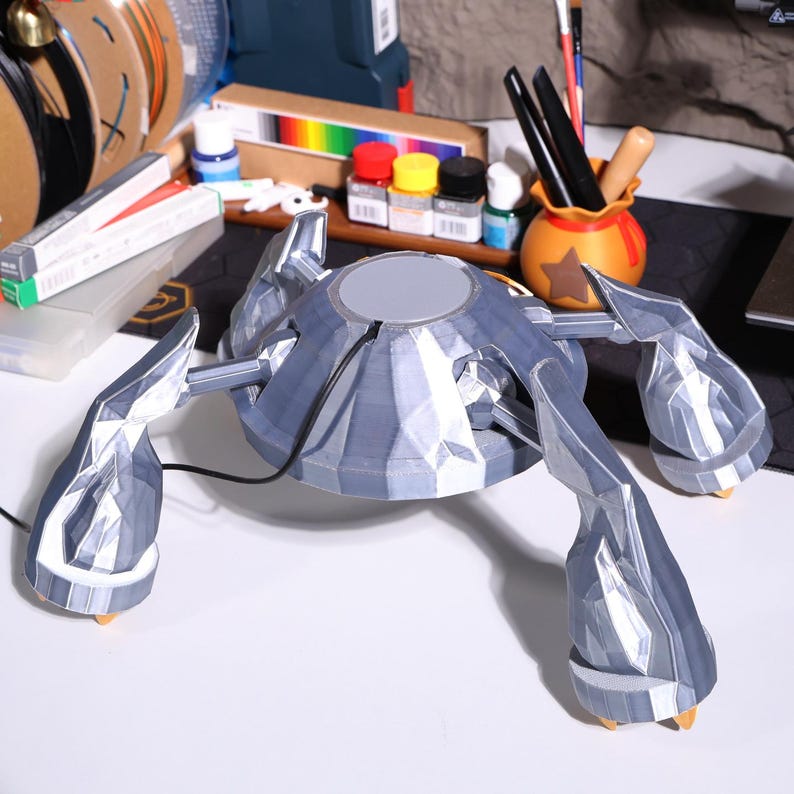 May include: A 3D printed, silver-colored, multi-legged robot with a round, flat top. The robot is sitting on a white surface.