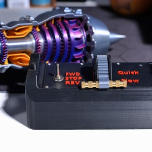 Op de afbeelding: Een zwart bedieningspaneel met een rode schakelaar met de tekst "FWD STOP REV", een grijze knop met een messing stang en een label met de tekst "Quick Slow".