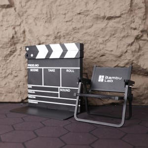 Artistic shot of the miniature film set from above, showing the layout and proportions of the chair and clapperboard together. This collectible is a tribute to the art of filmmaking and serves as a motivational symbol for creatives. Whether used as a photography prop, shelf accent, or personal keepsake, this set is an eye-catching piece for cinema fans, directors, and storytellers. The compact design ensures it fits perfectly in a variety of spaces, from home offices to studio desks.
