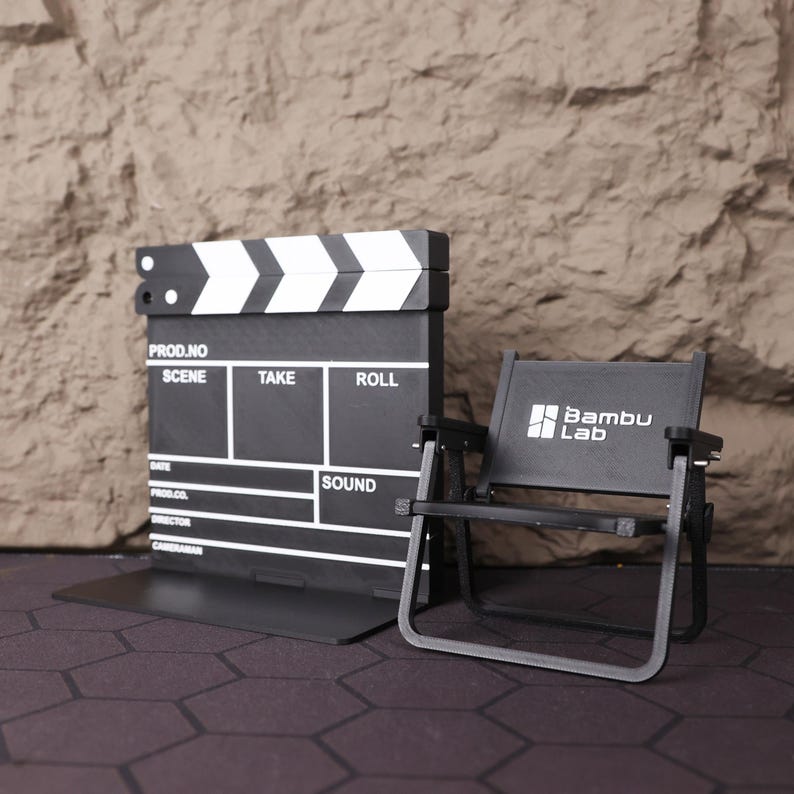 Artistic shot of the miniature film set from above, showing the layout and proportions of the chair and clapperboard together. This collectible is a tribute to the art of filmmaking and serves as a motivational symbol for creatives. Whether used as a photography prop, shelf accent, or personal keepsake, this set is an eye-catching piece for cinema fans, directors, and storytellers. The compact design ensures it fits perfectly in a variety of spaces, from home offices to studio desks.
