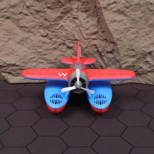 Op de afbeelding: Een rood en blauw speelgoed watervliegtuig met een witte propeller en de letter "W" op de romp. Het watervliegtuig heeft een rode vleugel en staart, met blauwe drijvers en een grijze motor.