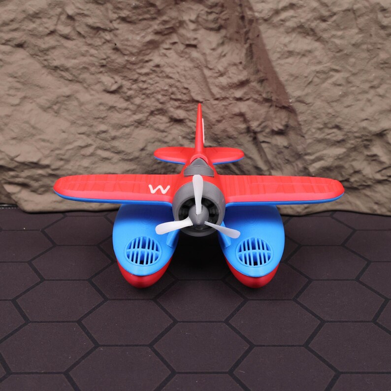 Op de afbeelding: Een rood en blauw speelgoed watervliegtuig met een witte propeller en de letter "W" op de romp. Het watervliegtuig heeft een rode vleugel en staart, met blauwe drijvers en een grijze motor.