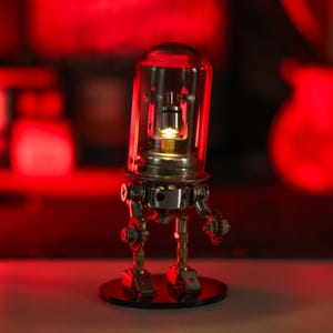 Puede incluir: Una pequeña lámpara robot plateada con una cabeza de vidrio transparente y una luz brillante en su interior. El robot está de pie sobre una base negra.