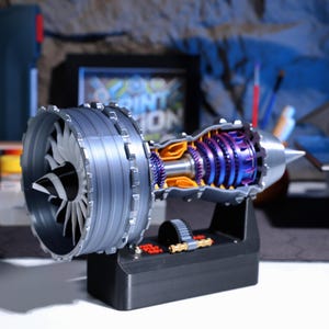 Op de afbeelding: Een gedetailleerd doorsnedemodel van een straalmotor, dat de interne werking van de turbine en compressor laat zien. Het model is geschilderd in tinten grijs, oranje en paars, en is gemonteerd op een zwarte basis.