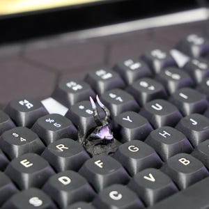 Puede incluir: Un teclado negro con una peque&ntilde;a figura de drag&oacute;n impresa en 3D, negra, sentada en las teclas F y G. El drag&oacute;n tiene cuernos blancos y una boca rosa.