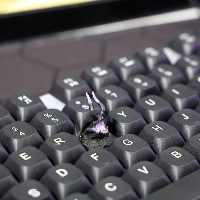 Puede incluir: Un teclado negro con una peque&ntilde;a figura de drag&oacute;n impresa en 3D, negra, sentada en las teclas F y G. El drag&oacute;n tiene cuernos blancos y una boca rosa.