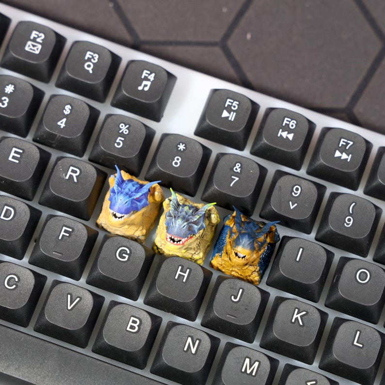 Puede incluir: Tres teclas de teclado con dise&ntilde;os de dragones. Las teclas son azules, amarillas y naranjas con detalles negros. Las teclas est&aacute;n en un teclado blanco.