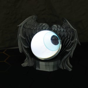 The LED crystal ball with custom magic eye is placed beside anime figures and gaming accessories, emphasizing its value as a holiday or birthday gift for him. Its crystal-clear orb and fiery visual inside enhance any gaming station, bookshelf, or nightstand, adding a touch of the supernatural with its gothic design and fantasy-themed projection that’s perfect for cosplay or room decoration.