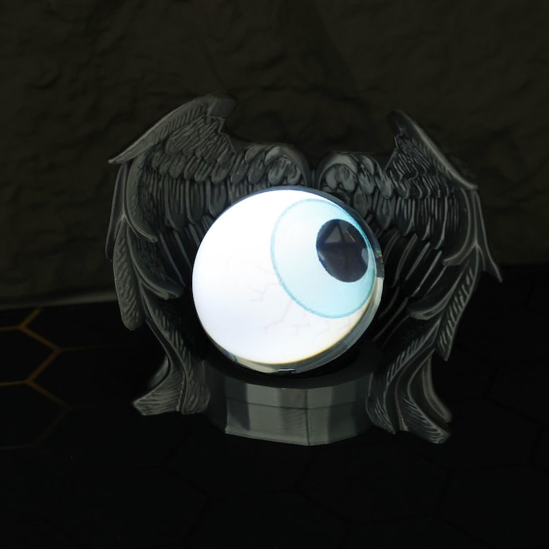 The LED crystal ball with custom magic eye is placed beside anime figures and gaming accessories, emphasizing its value as a holiday or birthday gift for him. Its crystal-clear orb and fiery visual inside enhance any gaming station, bookshelf, or nightstand, adding a touch of the supernatural with its gothic design and fantasy-themed projection that’s perfect for cosplay or room decoration.