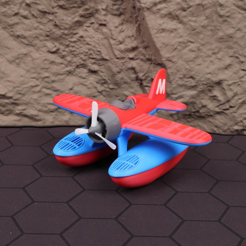 Op de afbeelding: Een rood en blauw speelgoed watervliegtuig met een witte propeller en de letter "M" op de staart. Het watervliegtuig heeft blauwe pontons en rode vleugels. Het speelgoed staat tegen een getextureerde, bruine achtergrond.