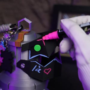 May include: A black and gray 3D printed helmet with a green and blue design. The helmet has a pink heart and the text "14" written on it. A person is using a pink marker to add details to the helmet.