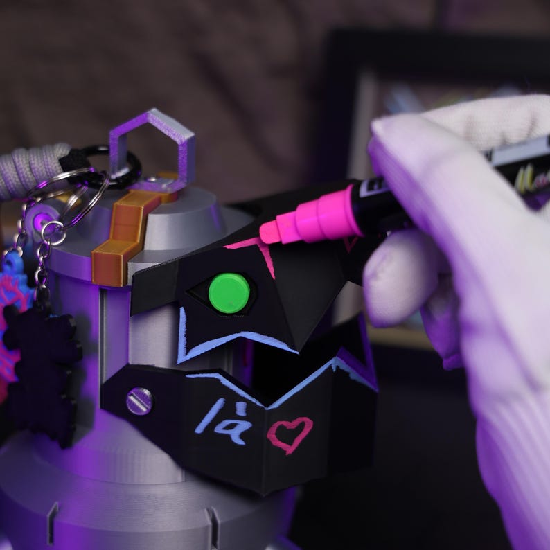 May include: A black and gray 3D printed helmet with a green and blue design. The helmet has a pink heart and the text "14" written on it. A person is using a pink marker to add details to the helmet.