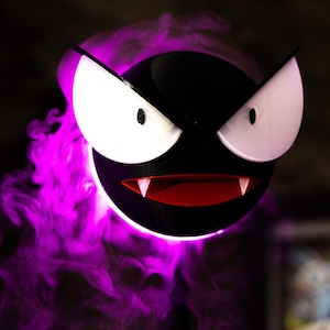 May include: A black, round, cartoon-like character with large white eyes, a red mouth, and two fangs. The character is surrounded by a cloud of purple smoke, with a dark background. The character is a popular design.