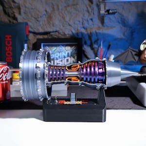 Op de afbeelding: Een gedetailleerd 3D-geprint model van een straalmotor, met een doorsnede die de interne componenten laat zien. Het model is geschilderd in tinten grijs, paars en oranje, en is gemonteerd op een zwarte basis.