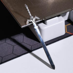 May include: A silver-colored sword with a detailed hilt and a sharp blade. The sword is resting on a black and gray textured surface.