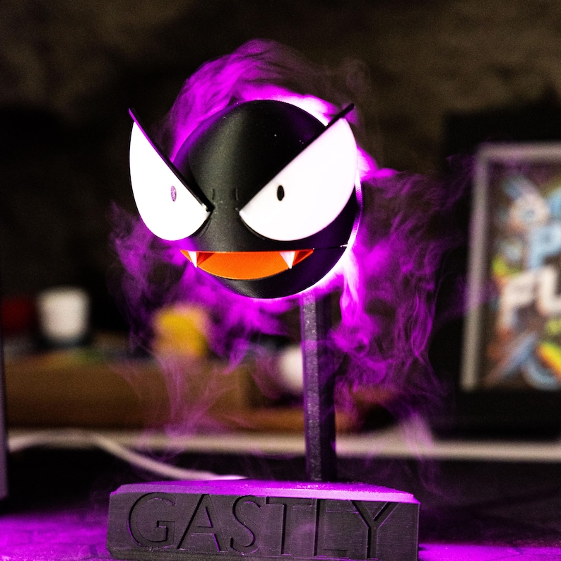 May include: A black Gastly figure with white eyes and an orange mouth, mounted on a stand. The base reads "GASTLY" in white letters. Purple smoke-like effects surround the figure, creating a spooky aesthetic.
