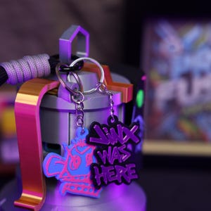 May include: A silver and gold grenade keychain with two purple and black charms. One charm says "Jinx was here" and the other is a cartoon character with pink, blue, and white details.