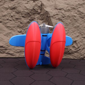 Op de afbeelding: Een 3D-geprint speelgoedvliegtuig met een blauwe romp, rode vleugels en een grijze propeller. De vleugels zijn ovaal van vorm en aan weerszijden van de romp geplaatst. De propeller heeft drie witte bladen. Het speelgoed is ontworpen om op een vintage vliegtuig te lijken.