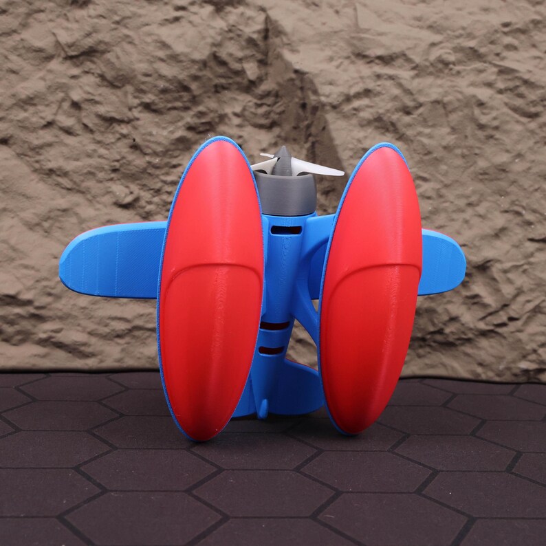Op de afbeelding: Een 3D-geprint speelgoedvliegtuig met een blauwe romp, rode vleugels en een grijze propeller. De vleugels zijn ovaal van vorm en aan weerszijden van de romp geplaatst. De propeller heeft drie witte bladen. Het speelgoed is ontworpen om op een vintage vliegtuig te lijken.