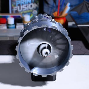 Op de afbeelding: Een close-up van een zilverkleurig 3D-geprint model van een straalmotor met een draaiende ventilator erin. De motor is gemonteerd op een zwarte basis.
