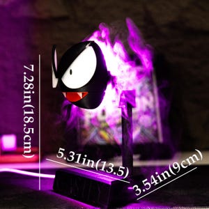 May include: A black and white cartoon character lamp with a red mouth and white eyes. The lamp is 7.28 inches (18.5cm) tall, 5.31 inches (13.5cm) wide, and 3.54 inches (9cm) deep. The lamp emits a purple glow and has a base that reads "GASTLY".