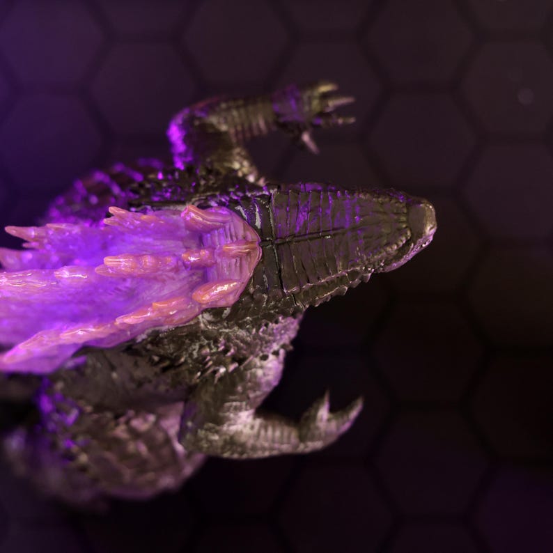 May include: A close-up of a black and silver monster figure with a purple and pink glowing dorsal fin. The figure is posed in a dynamic, action-ready stance.