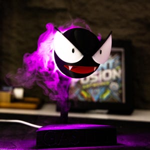 May include: A black and white cartoon character with a red mouth and fangs, and white eyes. The character is emitting purple smoke and is mounted on a black base. The base has the word "GAST" on it.