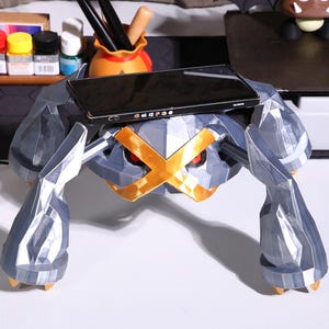 May include: A gray and silver 3D printed phone stand shaped like a Pokemon. The stand has a gold X across the front and a black smartphone is resting on top.