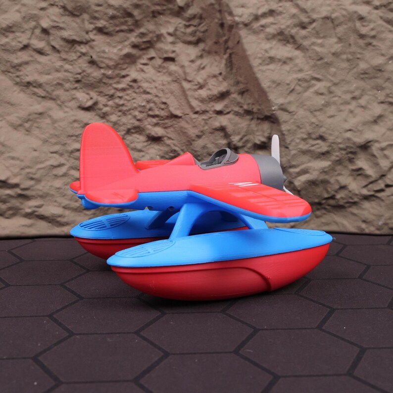 Op de afbeelding: Een rood en blauw speelgoed watervliegtuig met een propeller. Het vliegtuig heeft een rode romp en vleugels, met blauwe drijvers. De achtergrond is een getextureerd, bruin oppervlak.