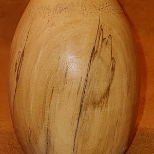 Può includere: Un uovo di legno di colore marrone chiaro con un motivo a venature del legno. L'uovo è liscio e ha una forma leggermente arrotondata.