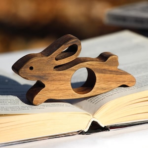 Peut inclure: Un anneau de serviette en bois en forme de lapin, sculpté dans une couleur brun clair, posé sur un livre ouvert. Le lapin est en position de course, les oreilles dressées.